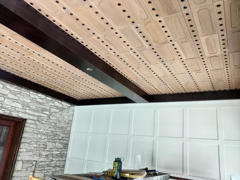 Op de afbeelding: Een plafond met lichtgekleurde rechthoekige houten panelen in een rasterpatroon, gescheiden door donkerbruine houten balken. De panelen hebben afgeronde verzonken centra en zijn voorzien van kleine, gelijkmatig verdeelde zwarte bevestigingen.