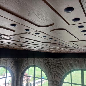 Op de afbeelding: Een lichtbruin houten plafond met een geometrisch patroon van rechthoekige panelen en verzonken donkere ronde accenten. Het plafond wordt ondersteund door een donkere horizontale balk boven drie boogramen met stenen kozijnen.