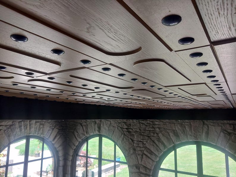 Op de afbeelding: Een lichtbruin houten plafond met een geometrisch patroon van rechthoekige panelen en verzonken donkere ronde accenten. Het plafond wordt ondersteund door een donkere horizontale balk boven drie boogramen met stenen kozijnen.