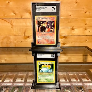 May include: Two graded Pokemon trading cards in protective cases. The top card is a holographic Japanese Charizard card graded 7. The bottom card is a holographic Venusaur card graded 8.
