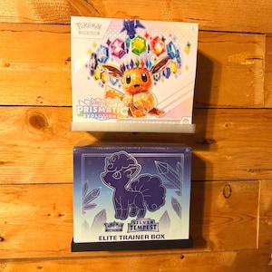 May include: Two Pokémon Elite Trainer Boxes on a wooden shelf. The top box features Eevee with colourful gems and the text "Scarlet & Violet Prismatic Evolutions." The bottom box has a purple Vulpix and reads "Silver Tempest Elite Trainer Box."