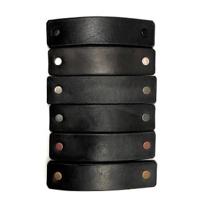 May include: Six black leather drawer pulls with different colored metal accents. The pulls are arranged vertically, with the top pull having a black metal accent, the second pull having a silver metal accent, the third pull having a silver metal accent, the fourth pull having a silver metal accent, the fifth pull having a copper metal accent, and the sixth pull having a gold metal accent.