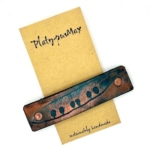 Puede incluir: Un brazalete de cuero marrón con un diseño de vaina de guisante en tono cobre y semillas de guisante verdes. El brazalete está hecho a mano y tiene el texto "PlatypusMax" y "hecho a mano de forma sostenible" en una etiqueta de papel marrón.