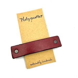 May include: A burgundy leather strap with two silver metal rivets. The strap is on top of a brown paper tag with the text "PlatypusMax" and "sustainably handmade".