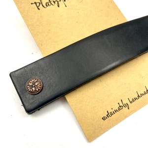 May include: A black leather strap with a copper-colored metal button featuring a floral design. The strap is lying on a brown paper tag with the text "sustainably handmade".