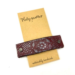 May include: A burgundy leather strap with a silver floral design and two silver metal rivets. The strap is on a brown paper tag with the text "PlatypusMax" and "sustainably handmade".