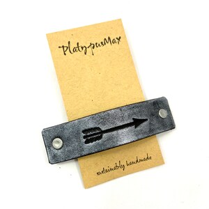 May include: A black leather bracelet with a silver arrow design. The bracelet is handmade and has the text "PlatypusMax" and "sustainably handmade" on a brown paper tag.