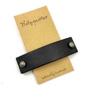 May include: Black leather strap with two silver metal studs. The strap is on a brown paper card with the text "PlatypusMax" and "sustainably handmade".
