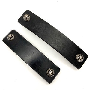 May include: Two black leather straps with silver metal accents. Each strap has two decorative metal pieces with a floral design.