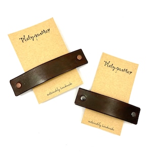 May include: Two brown leather hair clips with metal accents. The clips are on a brown paper background with the text "PlatypusMax" and "sustainably handmade".