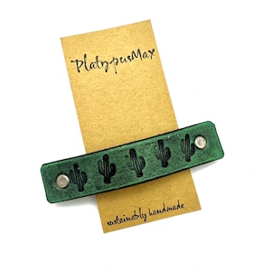 May include: Green leather wristlet with black cactus silhouettes and silver snaps. The wristlet is labeled "PlatypusMax" and "sustainably handmade".