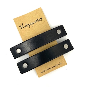 May include: Two black leather straps with silver metal buckles. The straps are on a brown paper background with the text "PlatypusMax" and "sustainably handmade".