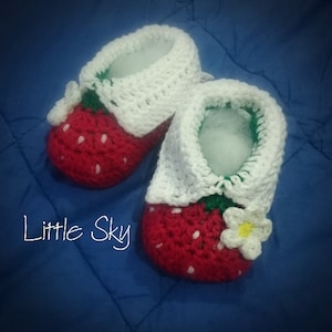 Könnte beinhalten: Ein Paar gehäkelte Babyschuhe in Form von Erdbeeren. Die Schuhe sind rot mit weißen Kernen und grünen Blättern. Jeder Schuh hat einen weißen gehäkelten Bund und eine weiße Blume mit gelber Mitte.