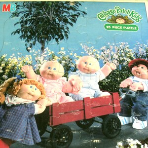 May include: A 25-piece Cabbage Patch Kids puzzle featuring dolls in a red wagon. The puzzle has a blue sky background with flowers and trees. The puzzle is for ages 3 to 7.