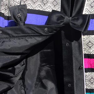 Op de afbeelding: Close-up van een zwarte jas met een satijnen afwerking, met strikdetails en drukknoopsluitingen. De jas heeft een patroon met zwarte, witte, paarse en roze strepen. De binnenvoering is zwart.