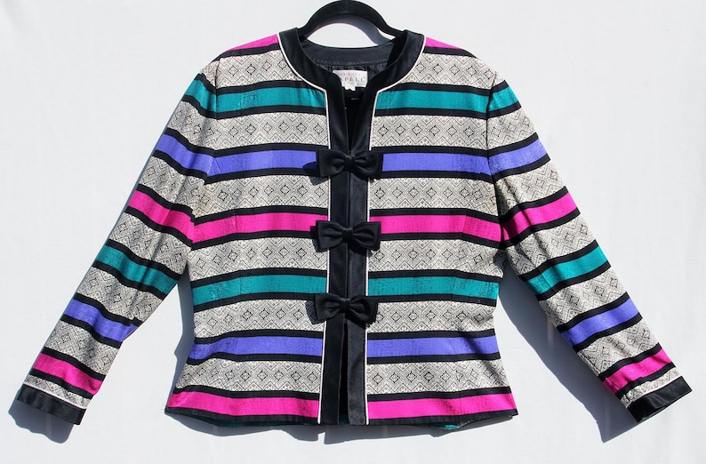 Op de afbeelding: Een vintage jasje met een opvallend streepjespatroon in teal, paars, roze en zilver. De jas heeft zwarte strikken aan de voorkant en zwarte bies. De mouwen zijn lang en de jas hangt aan een zwarte hanger.