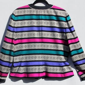 Op de afbeelding: Een vintage blazer met een opvallend streepjespatroon. De jas heeft horizontale banden in zwart, teal, paars, roze en een patroon in neutrale kleur. De kraag en manchetten zijn zwart met een witte bies. De blazer hangt aan een zwarte hanger.