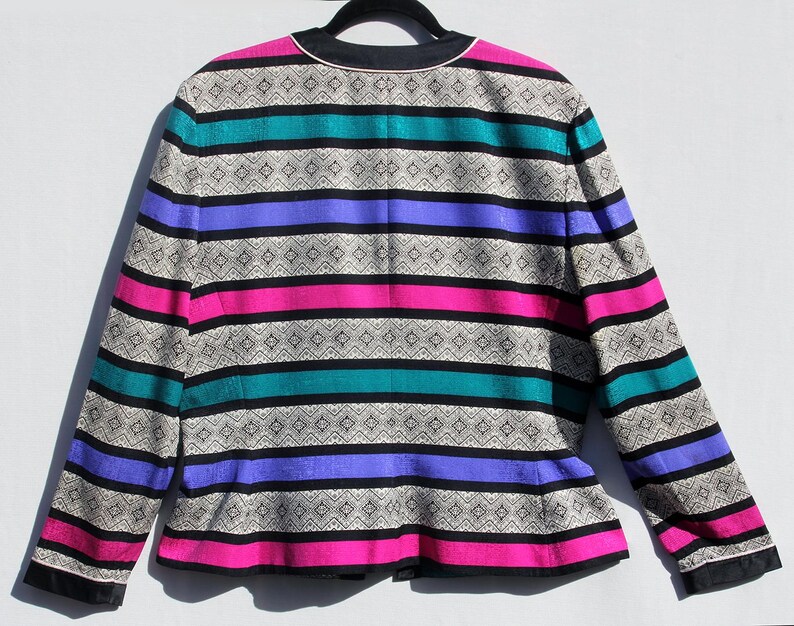Op de afbeelding: Een vintage blazer met een opvallend streepjespatroon. De jas heeft horizontale banden in zwart, teal, paars, roze en een patroon in neutrale kleur. De kraag en manchetten zijn zwart met een witte bies. De blazer hangt aan een zwarte hanger.