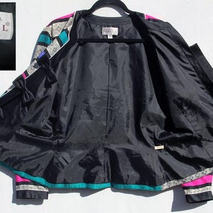 Op de afbeelding: Zwarte zijden jas met een levendig geometrisch patroon in turquoise, roze en wit. De jas heeft een zwarte voering, decoratieve strikken en het merklabel "Adrianna Papell". Maat 10.
