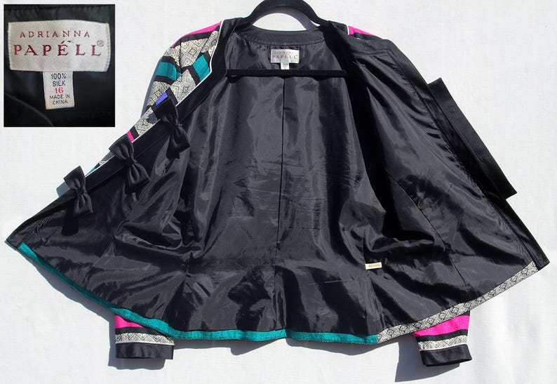 Op de afbeelding: Zwarte zijden jas met een levendig geometrisch patroon in turquoise, roze en wit. De jas heeft een zwarte voering, decoratieve strikken en het merklabel "Adrianna Papell". Maat 10.