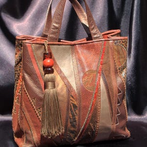 Puede incluir: Un bolso estructurado confeccionado con diversos paneles de cuero en tonos marrón y cobre con texturas variadas. El bolso cuenta con dos asas superiores y un accesorio decorativo de borla con dos cuentas grandes en tonos ámbar y marrón oscuro.