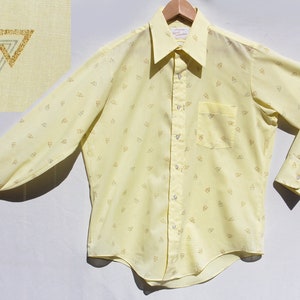 May include: A light yellow button-down shirt with a small triangle pattern. The shirt has long sleeves, a chest pocket, and a collar. The shirt is hanging on a wooden hanger. The upper left corner shows a close-up of the fabric with a logo.