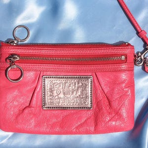 May include: A bright coral pink leather crossbody bag featuring a silver-toned metal plaque on the front with the embossed text Coach Poppy. The bag includes a zippered top closure, a front zippered pocket, silver hardware, and a detachable strap.