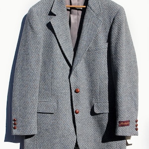 May include: A gray tweed blazer with brown buttons and a notched lapel. The jacket has two front pockets and a small label on the sleeve. The blazer is hanging on a wooden hanger.