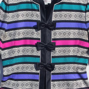 Op de afbeelding: Een kleurrijke jas met horizontale strepen in teal, paars, roze en zwart, geaccentueerd met een geometrisch patroon. De jas heeft een zwarte fluwelen bies en drie zwarte striksluitingen. Het label luidt "Dalkima Pelle".