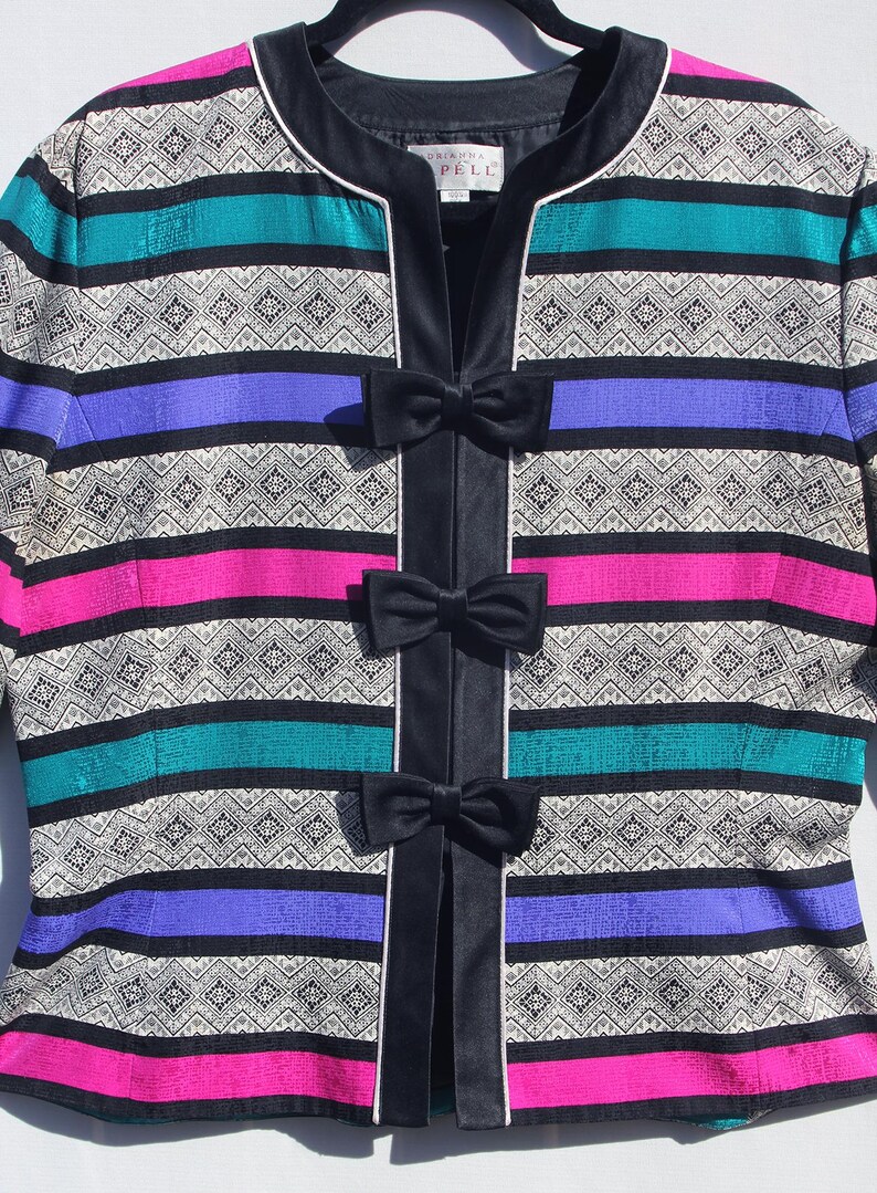 Op de afbeelding: Een kleurrijke jas met horizontale strepen in teal, paars, roze en zwart, geaccentueerd met een geometrisch patroon. De jas heeft een zwarte fluwelen bies en drie zwarte striksluitingen. Het label luidt "Dalkima Pelle".