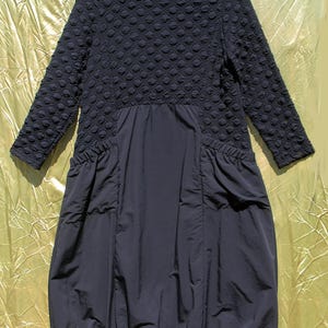 May include: A black dress with a textured, bubble-like pattern on the upper bodice and sleeves. The lower portion of the dress is a smooth, gathered fabric. The dress has a high, turtleneck collar and long sleeves. The dress is hanging on a hanger.