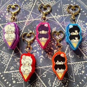 May include: A collection of keychains featuring a cartoon design of two characters stacked on top of each other, resembling ice cream cones. Each keychain has a gold-colored heart-shaped clasp and comes in various colors, including purple, pink, blue, red, and orange.