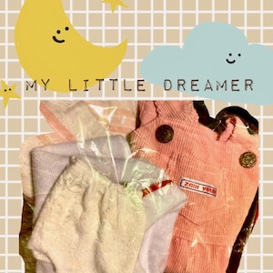 May include: A pink corduroy overall dress with gold-toned buttons and a small rectangular patch reading "HAPPY KIDZ" in red text, paired with a light purple fabric item, all encased in clear plastic packaging against a graphic background.