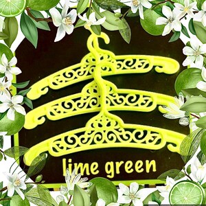 May include: Three lime green plastic clothes hangers featuring an intricate, openwork scroll pattern along the top arch. The hangers are stacked vertically against a dark background, with the text lime green printed in yellow at the bottom.