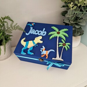 May include: Blue wooden box with a dinosaur, palm trees, and the name "Jacob" on the lid. The box has a blue ribbon tied around it.