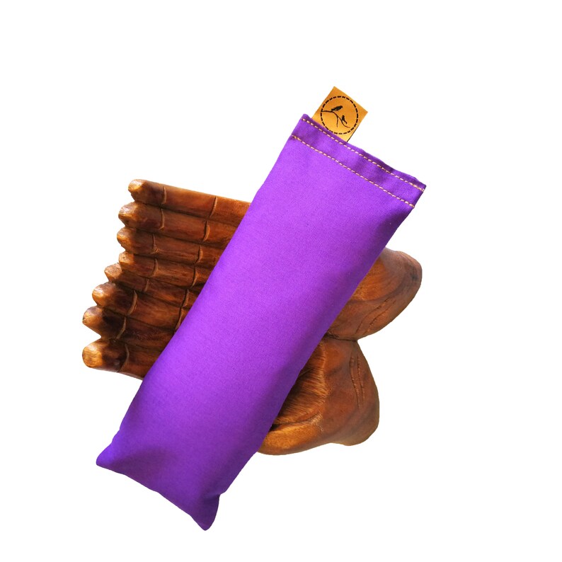 Puede incluir: Una vibrante almohada de aromaterapia morada descansa sobre un soporte de madera tallada. La almohada es rectangular, con un borde cosido. Se adjunta una peque&ntilde;a etiqueta con un logotipo. El soporte de madera tiene un dise&ntilde;o org&aacute;nico texturizado.