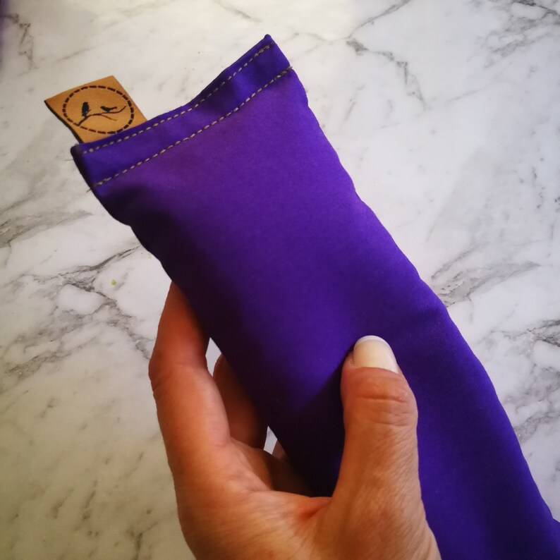 Puede incluir: Un vibrante juguete para gatos de color p&uacute;rpura con costuras doradas y una peque&ntilde;a etiqueta marr&oacute;n con un dise&ntilde;o de p&aacute;jaro. El juguete rectangular se sostiene en una mano, mostrando su color y textura. La etiqueta a&ntilde;ade un toque artesanal.