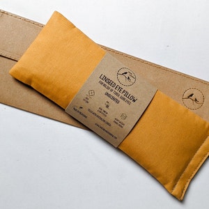 Puede incluir: Almohada para ojos de linaza de color mostaza con una funda de papel marr&oacute;n. La almohada lleva la etiqueta "Linseed Eye Pillow for Relief of Tired Sore Eyes Unscented". El embalaje incluye un peque&ntilde;o logotipo de p&aacute;jaro y detalles.