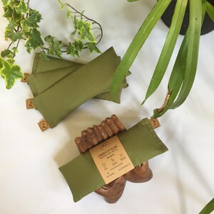 Puede incluir: Almohadas para los ojos de color verde oliva, de forma rectangular, se muestran sobre un soporte de madera y una superficie blanca. Cada almohada tiene una peque&ntilde;a etiqueta. La etiqueta dice "Linseed Eye Pillow" y "Unscented".