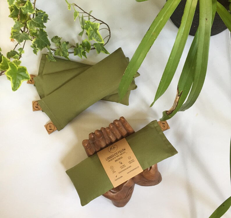 Puede incluir: Almohadas para los ojos de color verde oliva, de forma rectangular, se muestran sobre un soporte de madera y una superficie blanca. Cada almohada tiene una peque&ntilde;a etiqueta. La etiqueta dice "Linseed Eye Pillow" y "Unscented".