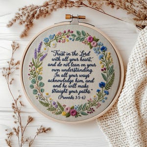 Op de afbeelding: Een ronde houten borduurring met lichtbeige stof en een bloemenkrans geborduurd in roze, blauwe, gele en groene draden. In het midden staat de donkerblauw geborduurde tekst Trust in the Lord with all your heart en de bijbelverwijzing.