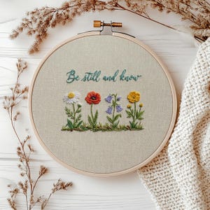 May include: A circular wooden embroidery hoop containing beige fabric with the hand-stitched teal text Be still and know above a row of embroidered wildflowers including a white daisy, a red poppy, blue bellflowers, and a yellow buttercup.