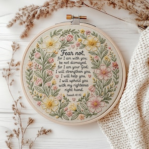 Peut inclure: Un tambour à broder circulaire en bois contenant un tissu blanc cassé avec une couronne florale brodée en fil rose, jaune et vert. Le centre présente un texte brodé en noir : Fear not, for I am with you; be not dismayed, for I am your God.