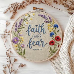 May include: A circular embroidery hoop containing beige fabric with the embroidered blue text 'faith OVER fear PSALM 34'. The text is framed by a colourful floral wreath featuring white daisies, purple lavender, yellow blossoms, blue cornflowers, and red poppies.