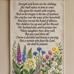 Peut inclure: Une tenture murale en tissu beige avec du texte brodé en noir tiré des Proverbes 31:25-30. La partie inférieure présente des broderies florales colorées, notamment des digitales violettes, des bleuets, du trèfle rose, des boutons d'or et des marguerites.