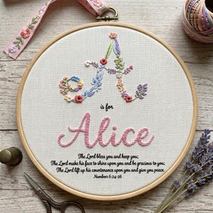 May include: A circular wooden embroidery hoop containing white fabric with a floral letter A, the name Alice in pink script, and the text The Lord bless you and keep you; The Lord make his face to shine upon you and be gracious to you; The Lord lift up his countenance upon you and give you peace. Numbers 6:24-26.