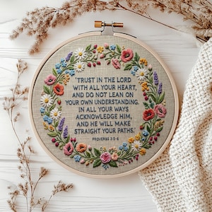 Könnte beinhalten: Ein runder hölzerner Stickrahmen mit beigem Stoff, der einen floralen Kranz aus rosa, gelben, blauen und grünen Fäden zeigt. In der Mitte steht der Text: TRUST IN THE LORD WITH ALL YOUR HEART, AND DO NOT LEAN ON YOUR OWN UNDERSTANDING. IN ALL YOUR WAYS ACKNOWLEDGE HIM, AND HE WILL MAKE STRAIGHT YOUR PATHS. PROVERBS 3:5-6.
