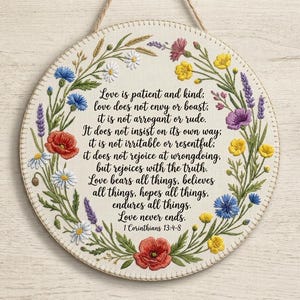 May include: A circular wall hanging featuring embroidered wildflowers in red, yellow, blue, and purple surrounding the black text of 1 Corinthians 13:4-8. The design is stitched onto an off-white fabric base with a stitched border and a hanging cord.