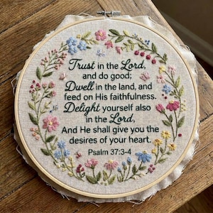 May include: A circular wooden embroidery hoop containing beige fabric with hand-stitched floral vines in pink, blue, and yellow. The center features black embroidered text: Trust in the Lord, and do good; Dwell in the land, and feed on His faithfulness. Delight yourself also in the Lord, And He shall give you the desires of your heart. Psalm 37:3-4.