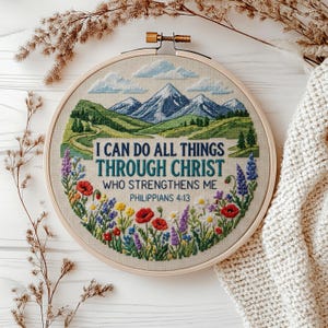 May include: A circular embroidery hoop containing a cross-stitch landscape of blue mountains, green hills, and a field of colorful wildflowers. The text reads: I CAN DO ALL THINGS THROUGH CHRIST WHO STRENGTHENS ME PHILIPPIANS 4:13 in blue lettering.